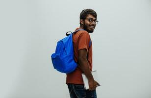 Indien étudiant avec bleu sac à dos, des lunettes et carnet posant sur gris Contexte. le concept de éducation et scolarité. temps à aller retour à école photo