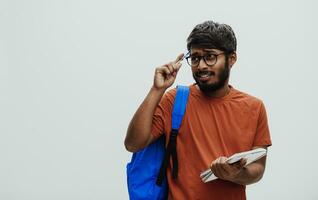 confus Indien étudiant avec bleu sac à dos, des lunettes et carnet posant sur gris Contexte. le concept de éducation et scolarité. temps à aller retour à école photo