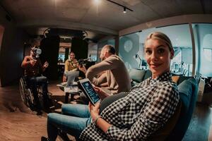 Enceinte femme séance sur le canapé de une moderne Bureau entouré par collègues et en utilisant une tablette photo