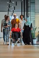 une diverse groupe de Jeune affaires gens en marchant une couloir dans le vitrée Bureau de une moderne Commencez, comprenant une la personne dans une fauteuil roulant et une femme portant une hijab, montrant une dynamique mélanger de innovation et unité. photo