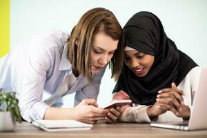 africain américain femme avec une hijab et une européen femme en utilisant une téléphone intelligent et portable dans leur Accueil Bureau photo