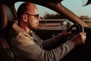 une homme avec une des lunettes de soleil conduite une voiture à le coucher du soleil. le concept de voiture Voyage photo