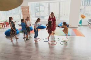 petit garderie école les enfants avec femelle prof sur sol à l'intérieur dans Salle de classe, Faire exercer. sauter plus de hula cerceau cercles Piste sur le sol. photo