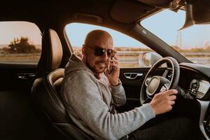 une homme avec une des lunettes de soleil conduite une voiture et parlant sur téléphone intelligent à le coucher du soleil. le concept de voiture Voyage photo