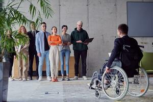 une groupe de diverse entrepreneurs recueillir dans une moderne Bureau à discuter affaires des idées et stratégies, tandis que une collègue dans une fauteuil roulant rejoint eux. photo