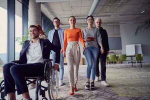une diverse groupe de les hommes d'affaires, comprenant une homme d'affaire dans fauteuil roulant, en toute confiance foulée ensemble par une moderne, spacieux bureau, incarnant collaboration, inclusivité, et force dans unité photo