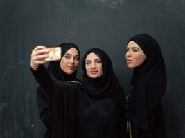 groupe de Jeune magnifique musulman femmes dans à la mode robe avec hijab en utilisant téléphone intelligent tandis que prise selfie image dans de face de noir Contexte photo