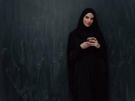 Jeune moderne musulman affaires femme en utilisant téléphone intelligent portant hijab vêtements dans de face de noir tableau noir photo