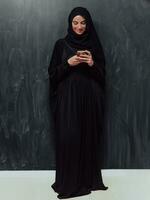 Jeune moderne musulman affaires femme en utilisant téléphone intelligent portant hijab vêtements dans de face de noir tableau noir photo