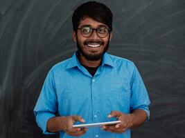 Indien souriant Jeune étudiant dans bleu chemise et des lunettes en utilisant tablette, téléphone intelligent et posant sur école tableau noir Contexte photo