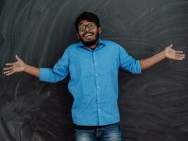 photo de un Indien homme portant une bleu chemise et des lunettes tandis que symboliquement montrant le sien vide les poches sur le Contexte de une école tableau noir.