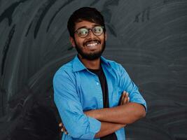 une Jeune Indien étudiant dans une bleu chemise avec des lunettes posant avec le sien bras franchi dans de face de le école tableau noir photo