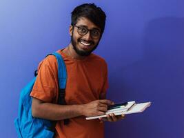 Indien étudiant avec bleu sac à dos, des lunettes et carnet posant sur violet Contexte. le concept de éducation et scolarité. temps à aller retour à école photo