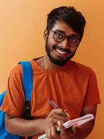 Indien étudiant avec bleu sac à dos, des lunettes et carnet posant sur Orange Contexte. le concept de éducation et scolarité. temps à aller retour à école photo