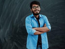 une Jeune Indien étudiant dans une bleu chemise avec des lunettes posant avec le sien bras franchi dans de face de le école tableau noir photo