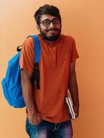 Indien étudiant avec bleu sac à dos, des lunettes et carnet posant sur Orange Contexte. le concept de éducation et scolarité. temps à aller retour à école photo