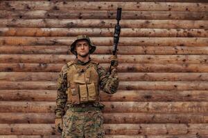 une soldat dans uniforme avec une fusil dans le sien main est permanent dans de face de une en bois mur. une soldat gardes le forêt base de le ennemi photo