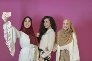 groupe de magnifique musulman femmes deux de leur dans à la mode robe avec hijab en utilisant mobile téléphone tandis que prise selfie image isolé sur rose Contexte représentant moderne Islam mode La technologie photo