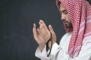 une Jeune arabe homme dans traditionnel vêtements fabrication une traditionnel prière à Dieu garde le sien mains dans prier geste dans de face de une noir Contexte photo