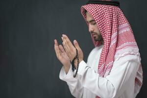 une Jeune arabe homme dans traditionnel vêtements fabrication une traditionnel prière à Dieu garde le sien mains dans prier geste dans de face de une noir Contexte photo