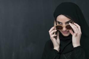 Jeune moderne musulman femme d'affaires en utilisant téléphone intelligent portant des lunettes de soleil et hijab vêtements dans de face de noir tableau noir photo