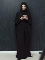 Jeune moderne musulman affaires femme en utilisant téléphone intelligent portant hijab vêtements dans de face de noir tableau noir photo