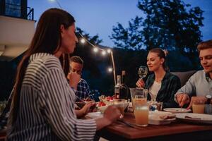 une groupe de Jeune diverse gens ayant dîner sur le terrasse de une moderne maison dans le soir. amusement pour copains et famille. fête de vacances, mariages avec barbecue. photo