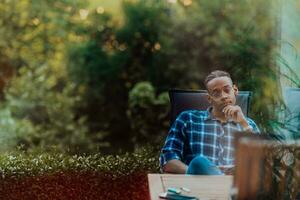 un africain américain homme séance dans de face de le maison sur une moderne terrasse et regarde pensivement à le caméra photo