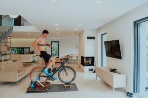 une homme équitation une triathlon bicyclette sur une machine simulation dans une moderne vivant chambre. formation pendant pandémie conditions. photo