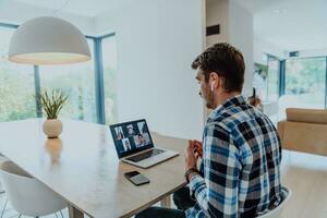 le homme séance à une table dans une moderne vivant chambre, avec écouteurs en utilisant une portable pour affaires vidéo discuter, conversation avec copains et divertissement photo