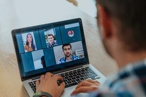 le homme séance à une table dans une moderne vivant chambre, avec écouteurs en utilisant une portable pour affaires vidéo discuter, conversation avec copains et divertissement photo