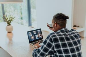 africain américain homme dans des lunettes séance à une table dans une moderne vivant chambre, en utilisant une portable et téléphone intelligent pour affaires vidéo discuter, conversation avec copains et divertissement photo