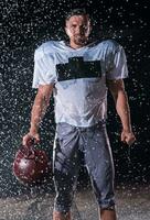 américain Football champ solitaire athlète guerrier permanent sur une champ détient le sien casque et prêt à jouer. joueur en train de préparer à courir, attaque et But atterrissage. pluvieux nuit avec spectaculaire brouillard, bleu lumière photo