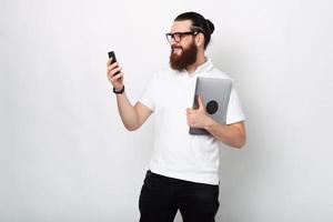 homme à la barbe en t-shirt blanc utilisant un smartphone et tenant un ordinateur portable