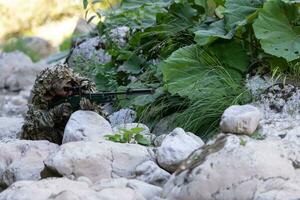 une militaire homme ou airsoft joueur dans une camouflage costume se faufiler le rivière et objectifs de une tireur d'élite fusil à le côté ou à cible. photo