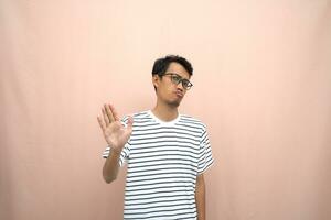 portrait de un asiatique homme dans des lunettes portant une décontractée rayé T-shirt. dénoter arrêt, traverser interdiction signe, interdire quelque chose, refuser,. beige Contexte. photo