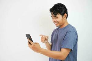 excité souriant ou content Jeune asiatique homme dans marine T-shirt en portant téléphone et montrer du doigt à téléphone intelligent avec doigt, isolé sur blanc Contexte photo