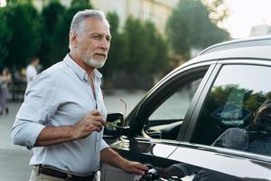 un homme âgé se tient près de sa voiture suv et tient des lunettes de soleil. photo