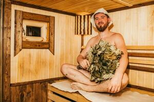 une homme avec une balai dans le sien main est relaxant dans le sauna photo