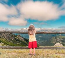 peu mignonne fille vu de derrière penché contre une en bois clôture est à la recherche à le montagnes photo