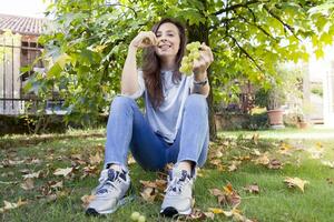 Jeune magnifique fille séance en dessous de une arbre en mangeant les raisins photo