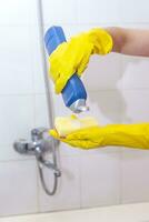 femme dans gants en mettant nettoyage chimie à le éponge et vouloir à nettoyer en haut le salle de bains photo