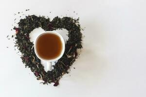 cœur forme fabriqué avec une mélange de une variété de séché thé feuilles et une tasse de noir thé photo