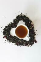 cœur forme fabriqué avec une mélange de une variété de séché thé feuilles et une tasse de noir thé photo