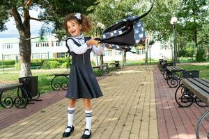 de bonne humeur marrant fille avec édenté sourire dans école uniforme avec blanc arcs dans école Cour lance le sac à dos. retour à école, septembre 1. content élève. primaire éducation, élémentaire classe. photo