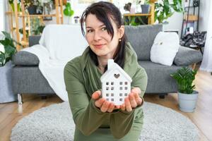 une content femme dans sa maison détient dans sa mains une miniature figure de une maison et clé dans le intérieur. rêver maison projet, réel biens achat, assurance, hypothèque, louer, réservation photo