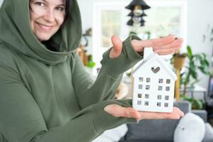 une content femme dans sa maison détient dans sa mains une miniature figure de une maison et clé dans le intérieur. rêver maison projet, réel biens achat, assurance, hypothèque, louer, réservation photo