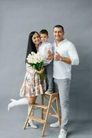 portrait de une content Jeune élégant famille avec une petit enfant sur une gris Contexte. de la mère journée avec fleurs photo