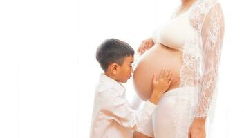 Jeune garçon est embrasser le Enceinte femme isolé sur blanc Contexte photo