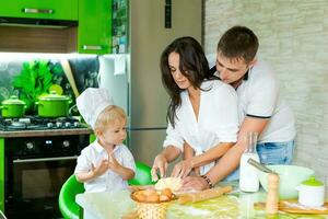 content famille maman et peu fils et papa sont en train de préparer pâte dans cuisine à tableau. des produits pour pâte sont sur table photo